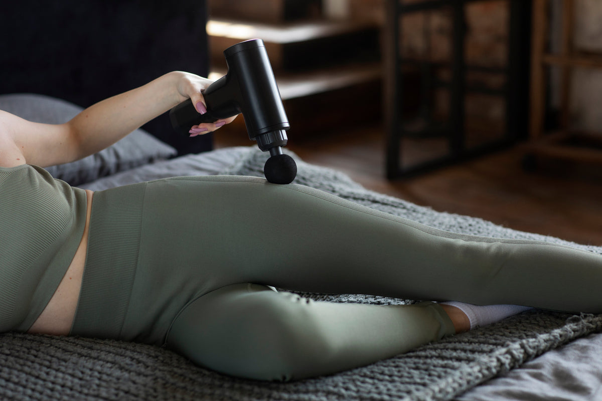 Woman lying on a bed using a massage gun on her glutes for muscle recovery